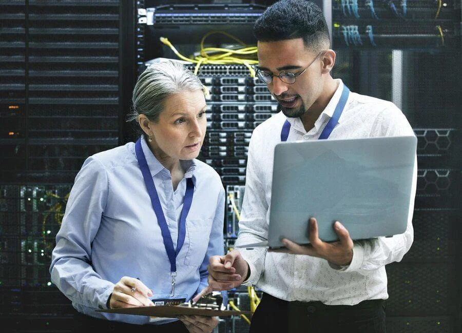 Two IT professionals discussing in front of server racks, one holding a laptop and the other a clipboard.
